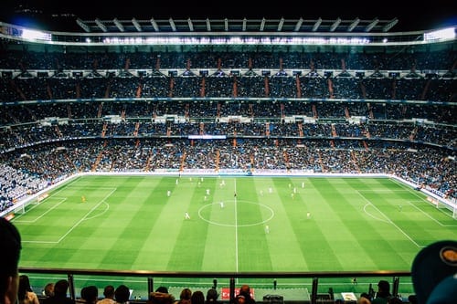 A large, semi-covered sports stadium in which a soccer game is in progress. Approximately five tiers high, filled to capacity, pitch is a verdant green.