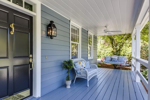 Covered verandah on the front of a blue wooden house. The front door is dark blue, the verandah ceiling and framework are white as are the window frames. A white wicker couch and plant is beside the front door, which had a brass knocker.