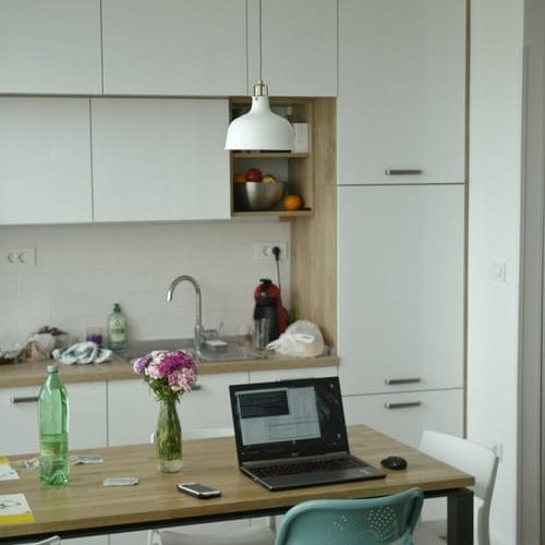 A small kitchen and a dining room table, with modern, fresh white decor. An open laptop is sitting on the table, demonstrating that this person is working from home. A bottle of water, a vase of flowers and a mobile phone are also on the desk.