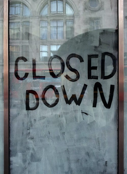 Dusty window, with "CLOSED DOWN" written by hand in black. You can't see into the window due to the dust/dirt. However you can see a reflection of the buildings across the road on the glass. They are older, multi-storied stone structures, and a red double-decker bus is travelling along in front of the building.