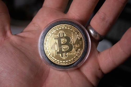 Looking down on the palm of man's left hand containing a bitcoin. The coin is sitting flat, it is black and gold. The man is wearing a plain gold wedding band.