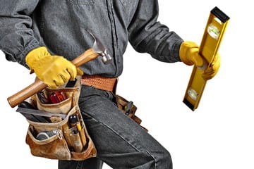 Side-on view of a construction worker from just below his shoulders to his knees. He is wearing a dark, grey shirt and black jeans. He has on a brown tool belt, and is carrying a hammer and yellow level.