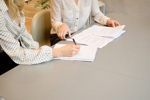 Two women sitting side by side at large table, one reading through a contract before signing it. The other is taking notes. Both are wearing spotted blouses, and are in an environment.