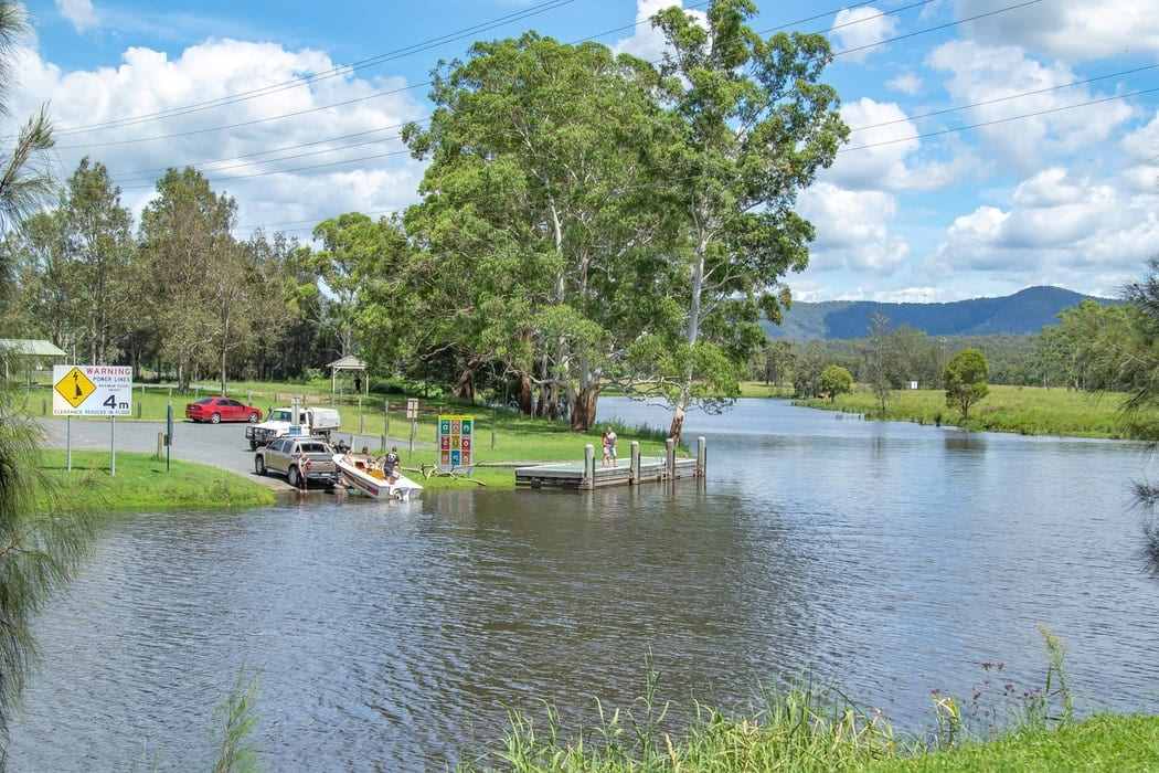 Rural scene of a boat being launched at a river. A car is backing a small runabout down the boat ramp. In the background are very large trees and mountain ranges. The water is slightly brownish in colour and rippling. A small wooden jetty sits to the side of the boatramp.