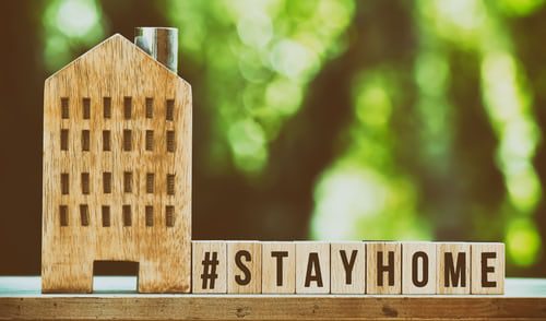 A small, wooden model of an apartment building sitting on a window ledge. Beside the model are the words "#STAYATHOME" spelt out in small wooden building blocks. Outside the window, in the background, are blurred, green trees.