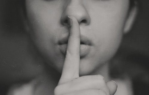 Black and white photo of a woman holding her finger upright in front of pursed lips indicating to keep a secret or to "sshhh". The photo does not show her eyes, it finishes just above her nose.