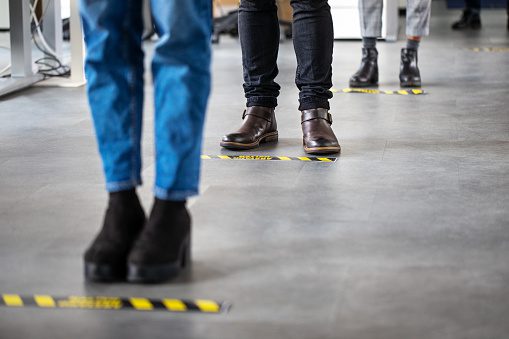 Four people, standing in a line on a grey polished floor. The people are shown from the knees down, and are evenly spaced one behind he other. Each is behind a fine yellow and black line on the concrete, indicating where they should stand.