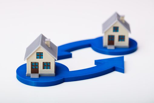 Small, plastic models of two houses sitting side by side on a white flat surface, a few inches high. They are predominantly cream and grey. Each has a blue arrow pointing to the other.