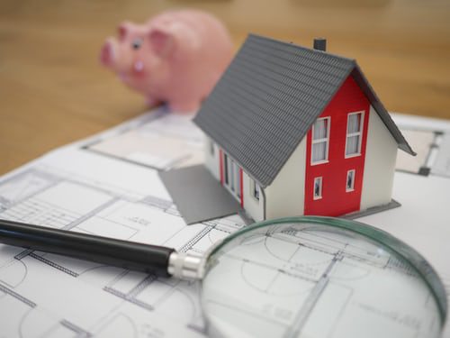 Small, plastic model of a red, grey and white house, sitting on a set of house plans on a desk. In front of the house is a black and chrome magnifying glass, behind the house is a pink, ceramic piggy bank.