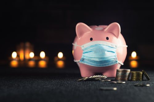 Small, pink ceramic piggy bank standing behind a small pile of coins, some of which have fallen over, and some are just tilted on an angle. Behind the pig are some blurred candle flames showing in the dark. The pig is wearing a blue and white paper covid mask.