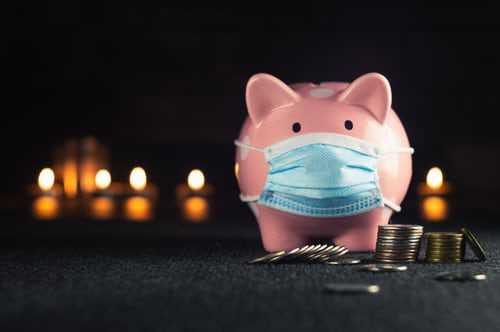 Small, pink ceramic piggy bank standing behind a small pile of coins, some of which have fallen over, and some are just tilted on an angle. Behind the pig are some blurred candle flames glowing in the dark. The pig is wearing a blue and white paper covid mask.