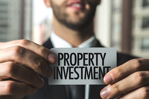 Close up shot of a man holding a small white sign with Property Investment on it. The front of the shot features the man's hands holding the sign between his fingers. The back of the shot is blurred and shows the man in the background wearing a suit.
