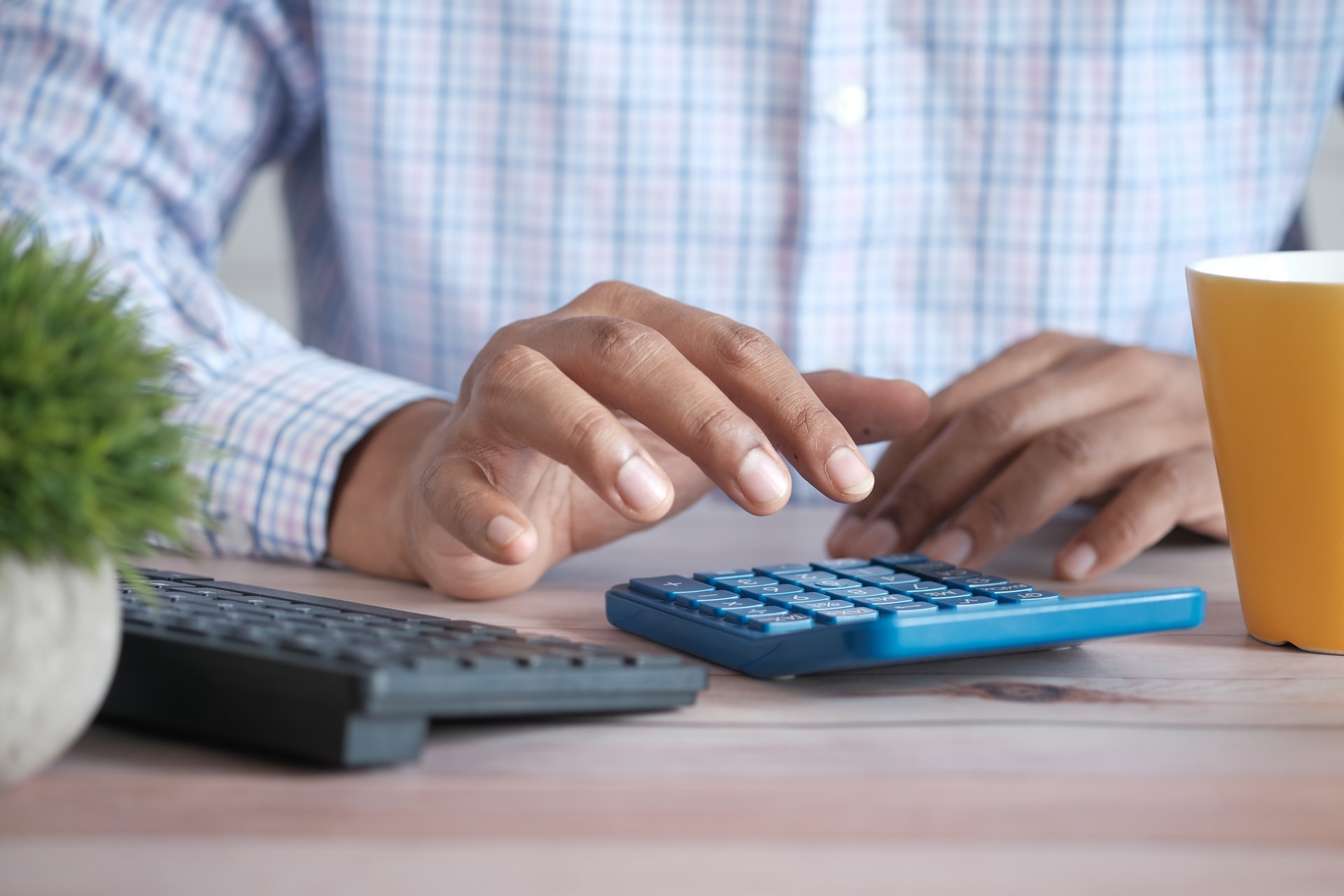 Man sitting at a desk using a blue calculator, which is sitting beside a black computer keyboard. He is wearing a pink, white and blue checked shirt, and has a orange coffee mug in front of him.