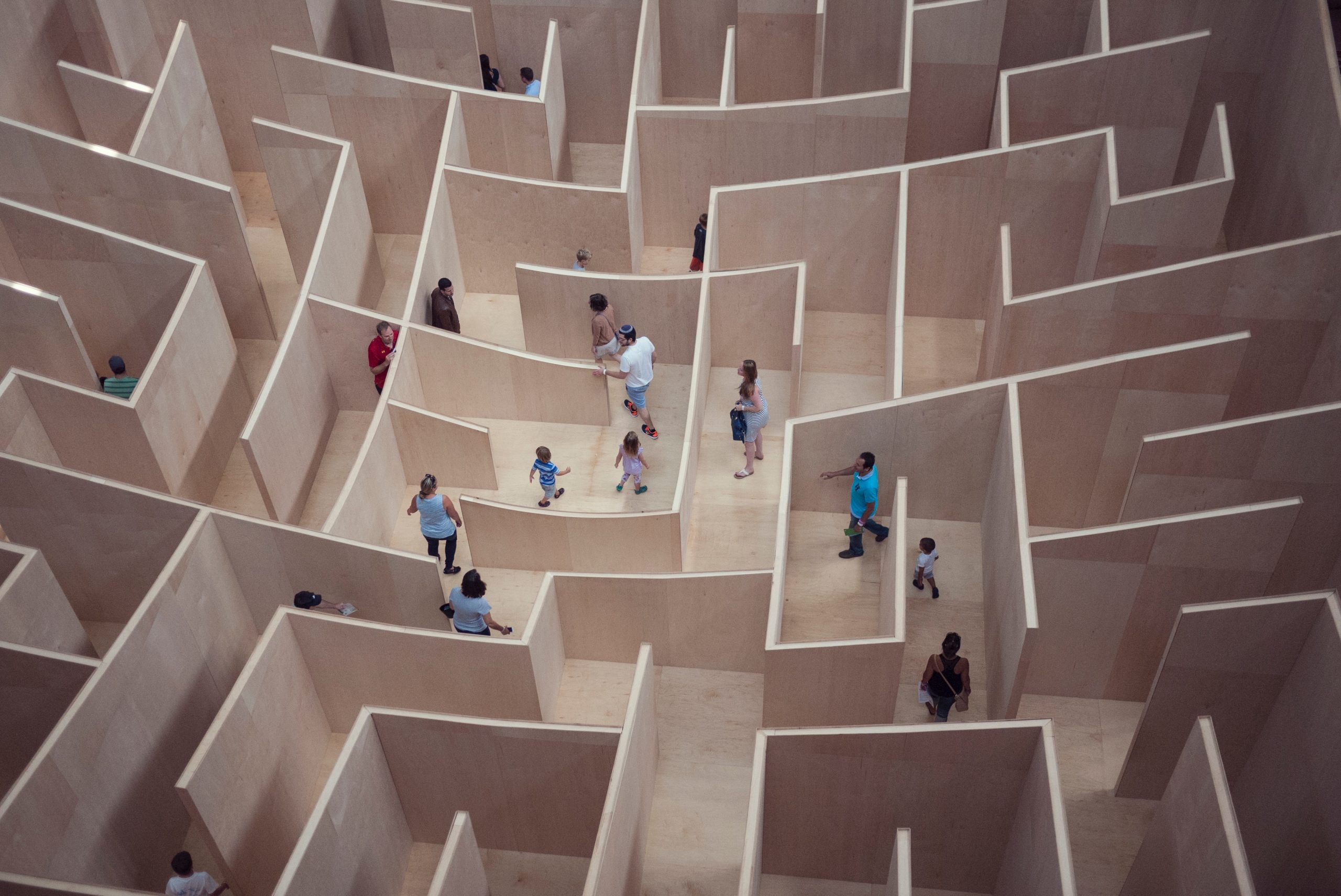 Bird's eye view of a wooden maze with approximately 18 people inside. Some adults, some children. Wood is of a light colour, some of the wooden walls are low and can be seen over. Others are much taller than the people.