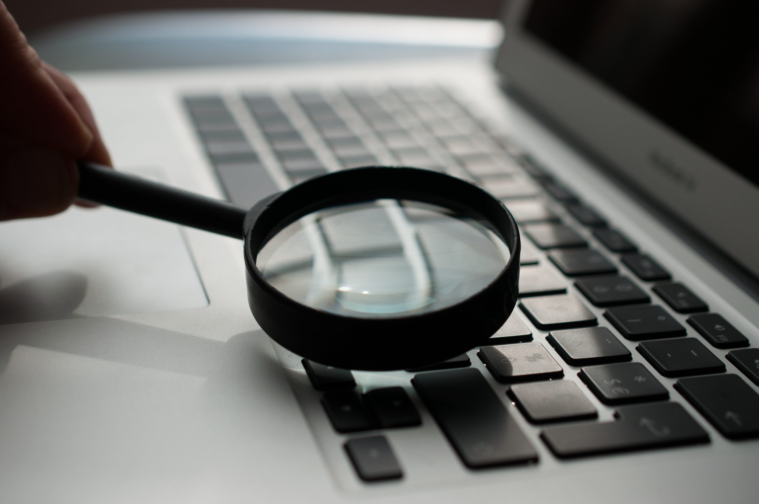 Person's hand in top left of photo, placing a plain black magnifying glass on to the keyboard of a computer screen. Everything in the photo is blurred apart from the magnifying glass.