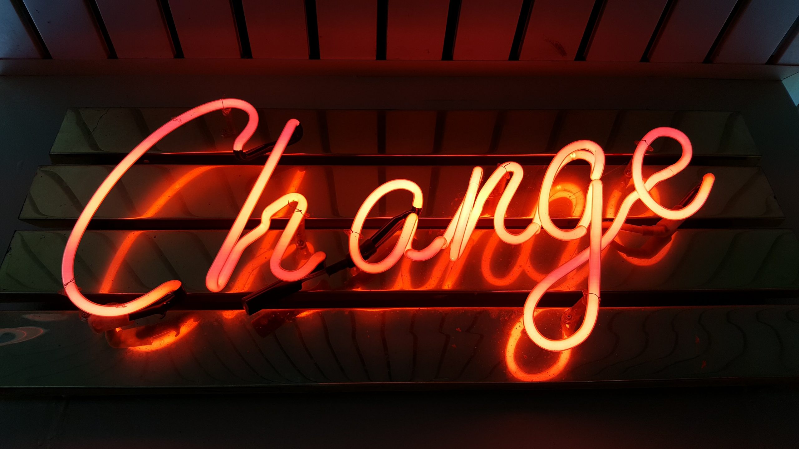 Bright orange neon sign spelling out "Change" with a capital C and then all lower case. Appears to hang from ceiling in dimly lit room.