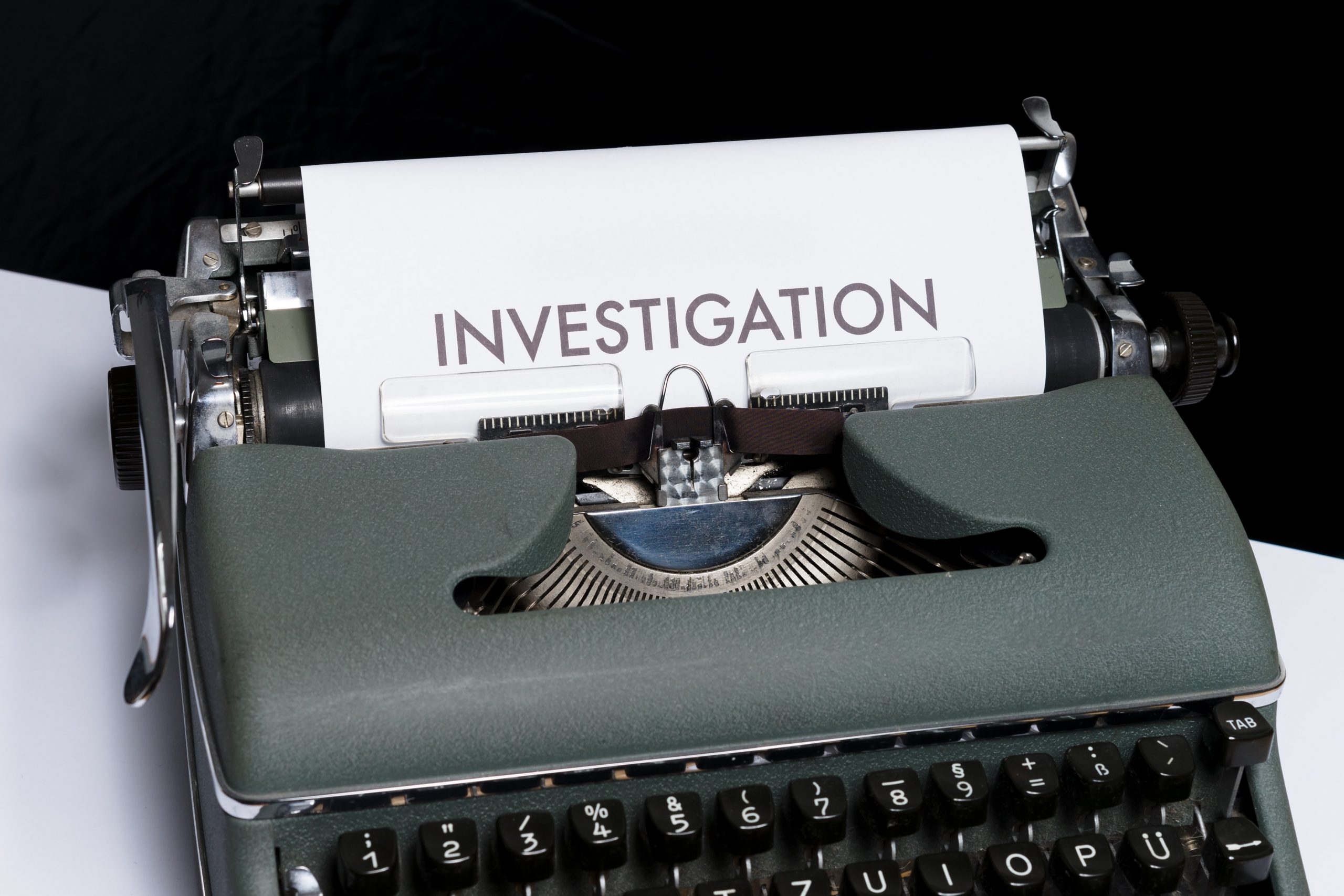 Old fashioned green, metal typewriter sitting on desk. White paper in the typewriter has the heading "Investigation" written at the top.