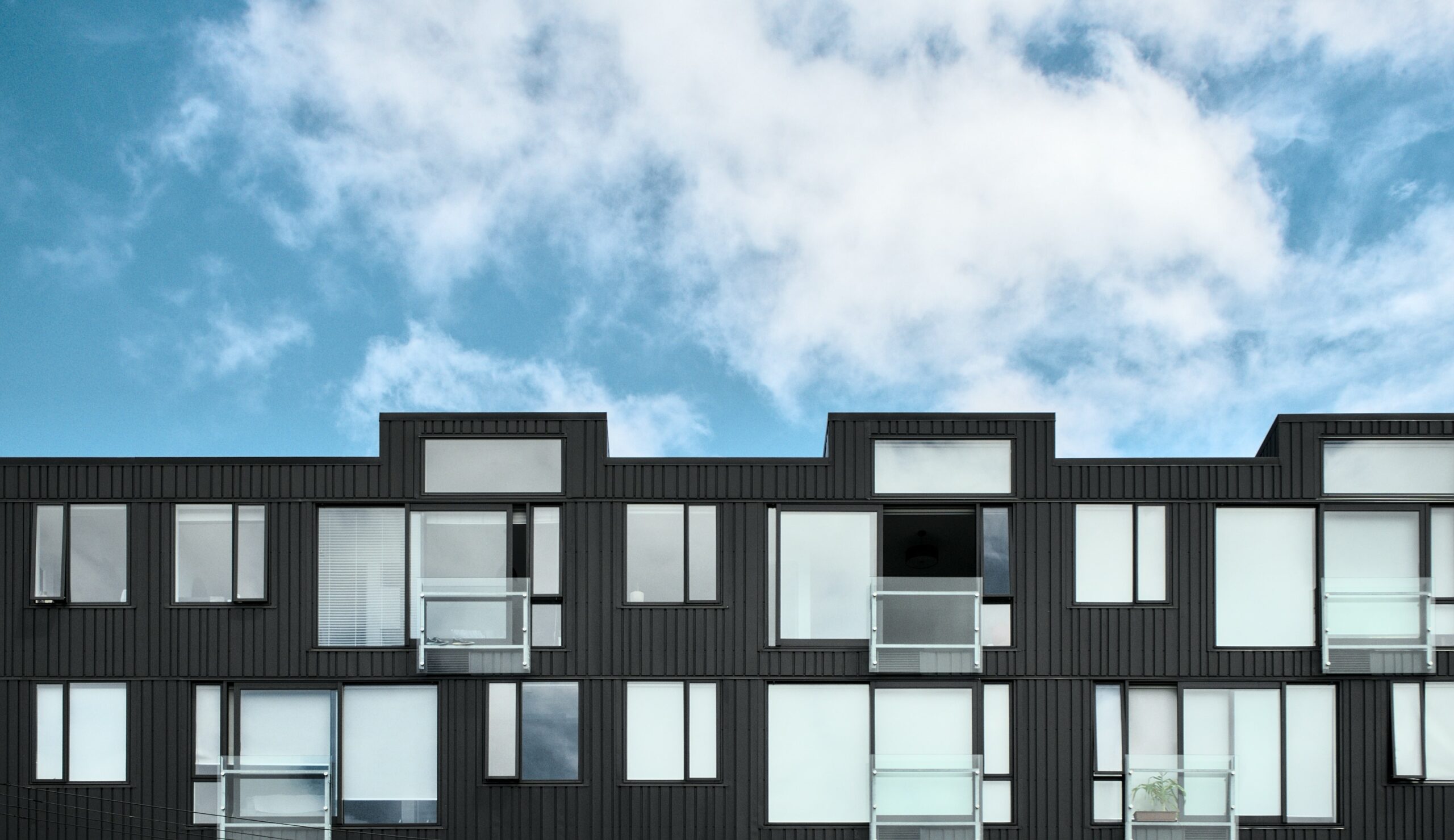 Horizontal view of black and white apartment block. Top two levels are pictured, with large windows, small balconies and white blinds.