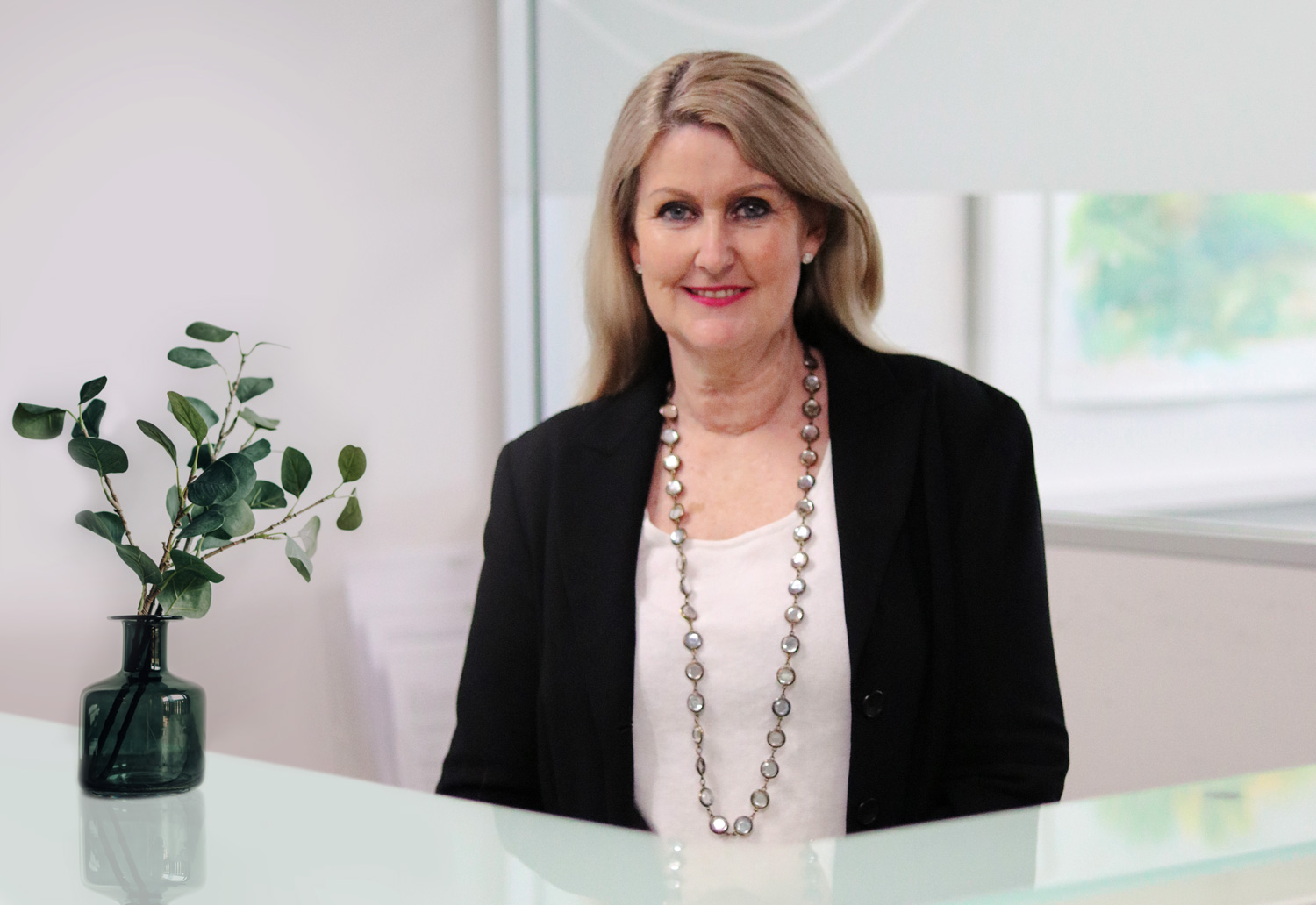 Practice Manager standing at reception. Wearing a black jacket, white top and silver necklace. Reception desk has a small green indoor plant sitting on it.
