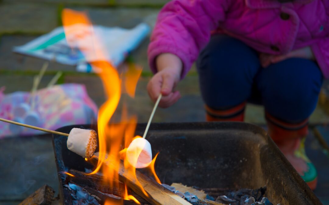 Child crouching down over an open fire, toasting a marshmallow on a wooden skewer. Another person, not in the picture, is also holding a skewer in the flames. Child is wearing a pink/purple jacket and multi-coloured gumboots.