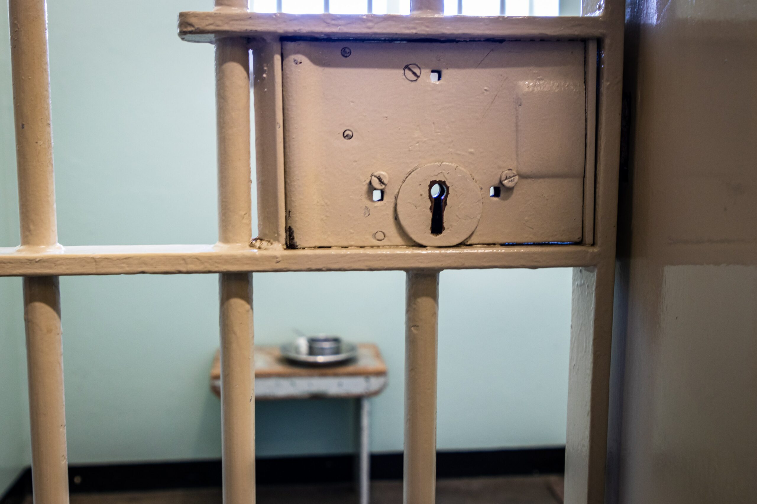 Close up of beige painted metal barred door of prison cell. Inside is a small table with a metal plate and cup sitting on it.