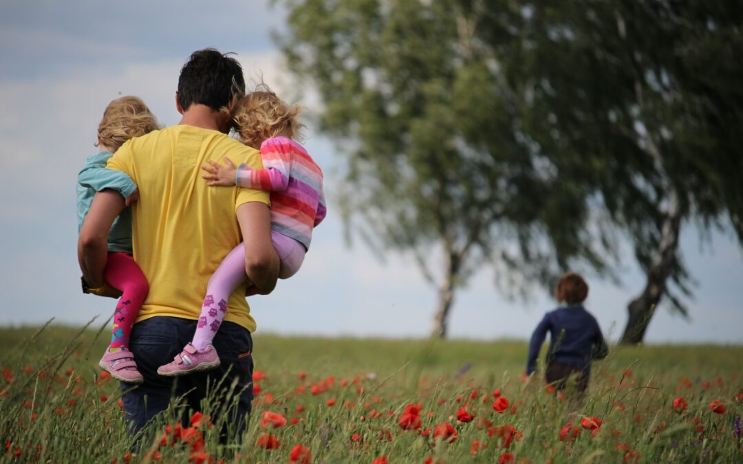 Man carrying two little girls walking through poppy field. Young boy is running ahead of them towards a group of trees.