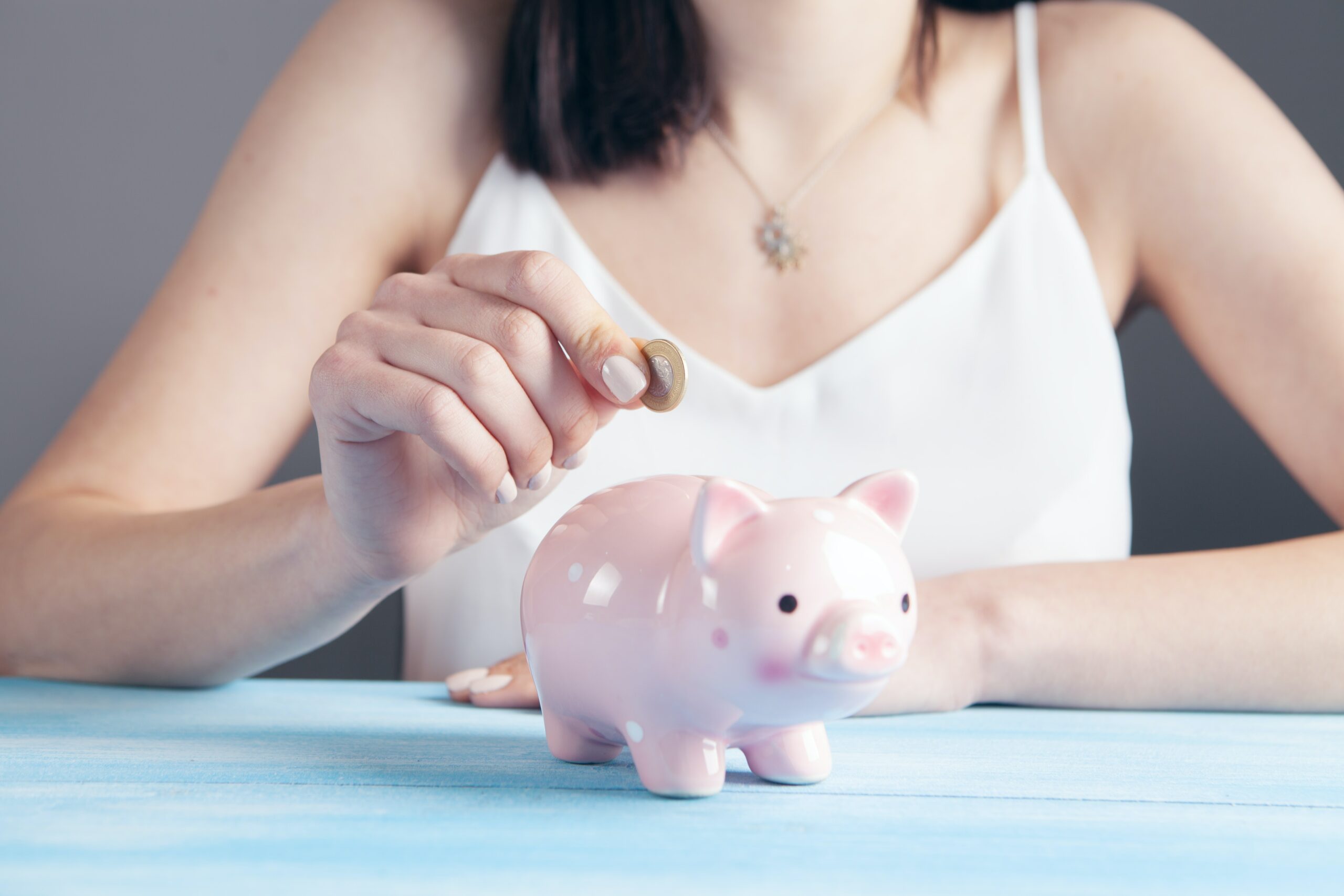 Woman putting gold coin into pink ceramic piggy bank. She is wearing a white camisole top, has dark hair and pink painted nails.