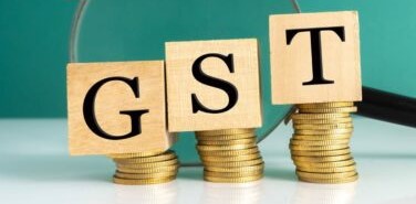Three small, wooden building blocks in a line, each with a letter on the front, spelling GST. Under each block is a small stack of gold coins. Each stack is a different height, ascending from the lowest at G, up to the highest at T. Behind the blocks is a black and chrome magnifying glass lying on its side.
