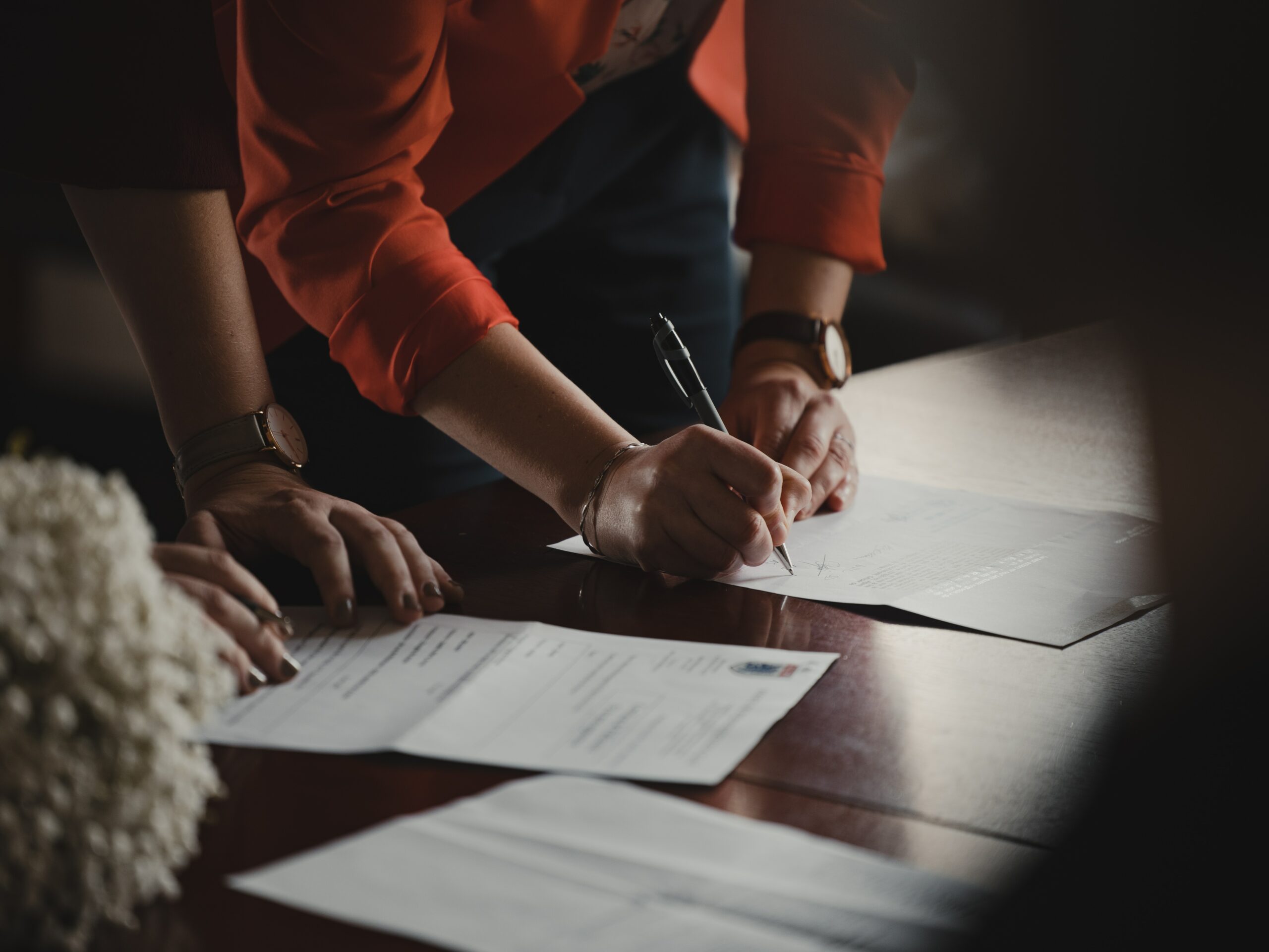 Two women leaning on a wooden desk appearing to sign a contract. Both are wearing watches and appear to be in work attire.