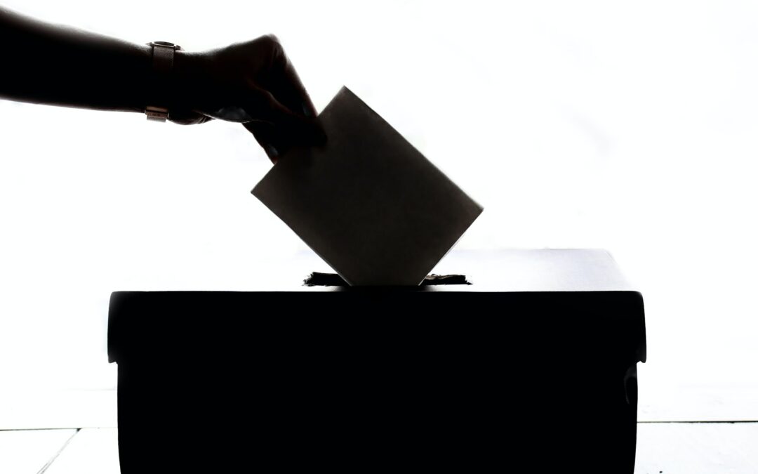 Shadowed photo showing woman's forearm and hand holding a voting slip and dropping it into a voting box.