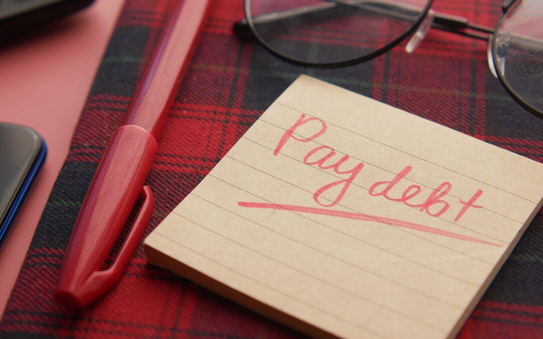 Small yellow Post It note pad, sitting on red and black tartan cloth, beside which is a pair of glasses and a red pen. "Pay debt" is written in red ink.