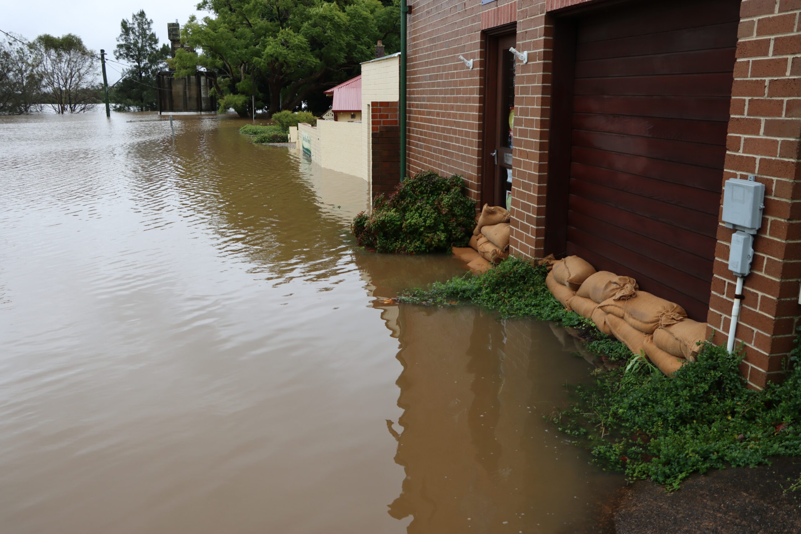 Wide rural street flooded with brown water, several feet high. Brick building at front has sandbags in doorways and greenery/shrubs under water.