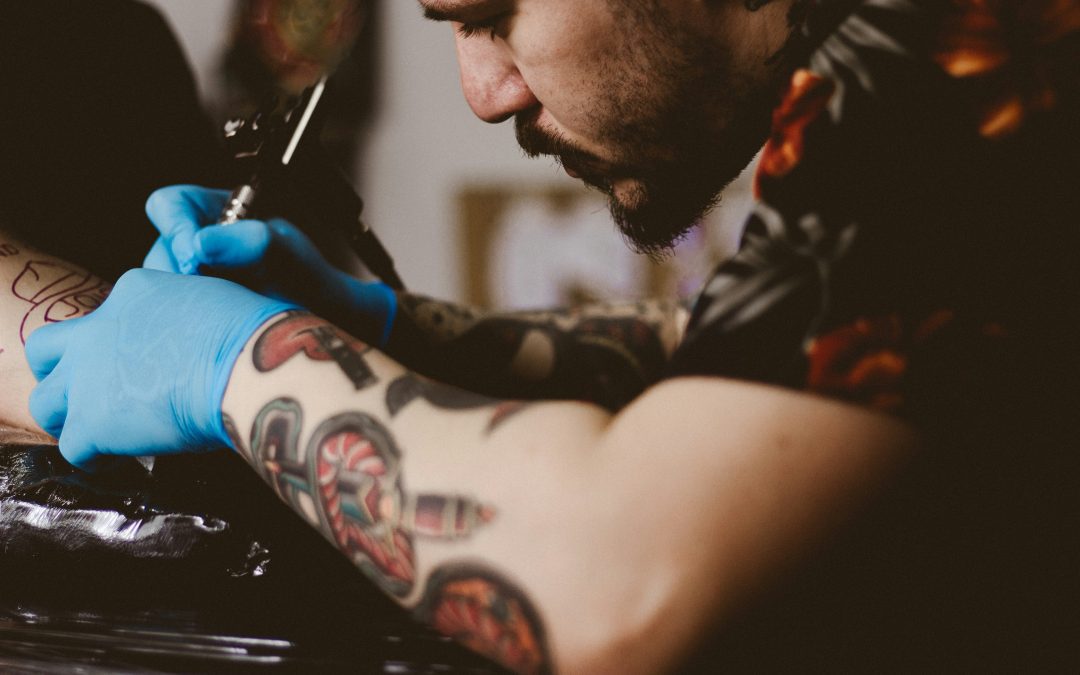 Close up of a male tattooist, inking a client. The tattooist himself has his forearms heavily tattooed.