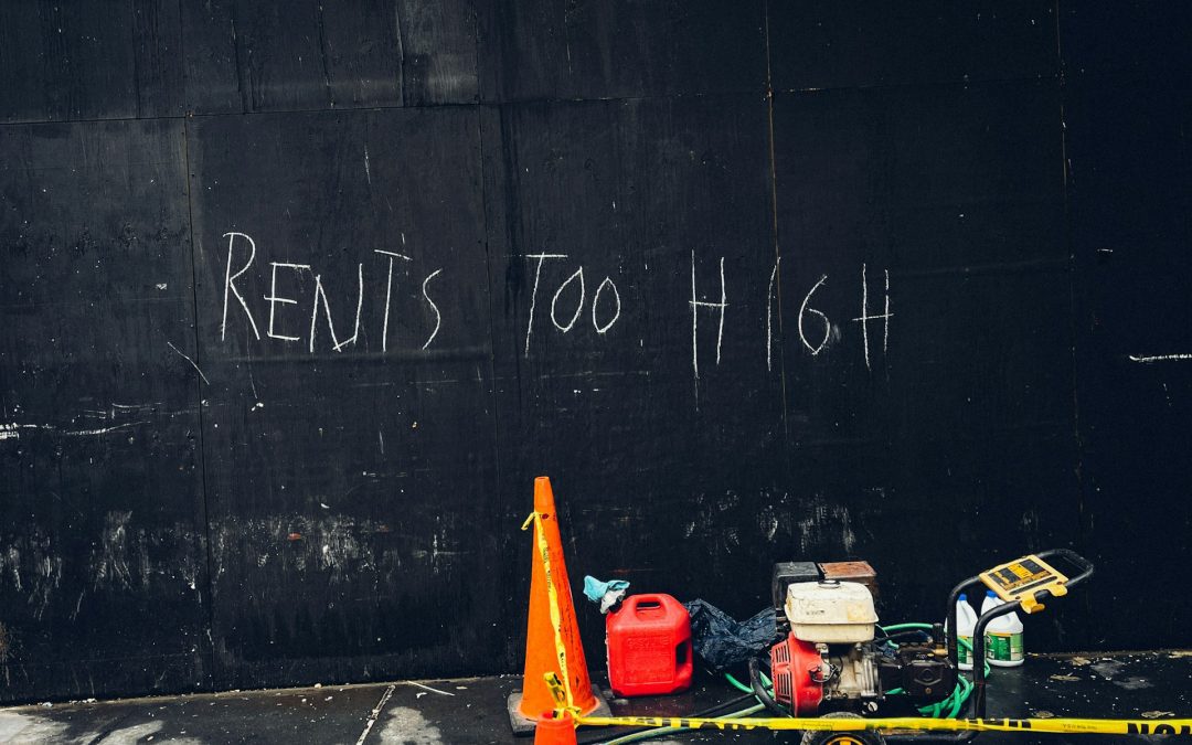 "Rents too high" written in white chalk on a black, wall. Underneath the text, on a concrete footpath, is a water-blaster, orange cones and cleaning equipment, suggesting the graffiti is about to be cleaned off.