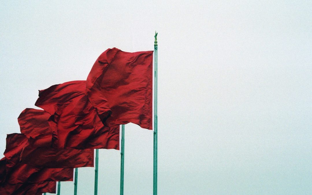 A row of six red flags on metal flagpoles, ascending in height from left to right., starting at the bottom left of the shot and moving to the top right. They are blowing in the wind, against an almost clear, white sky.