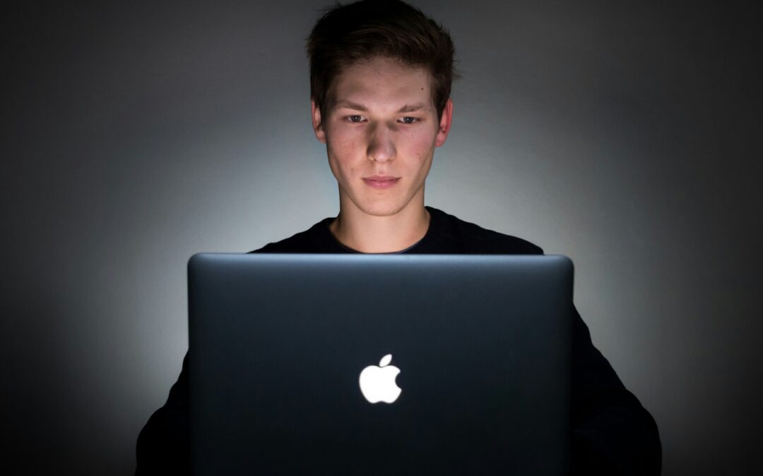 A young man sitting facing us working on an Apple computer on the desk in front of him. Shaded, dark background, he is also dressed in black.