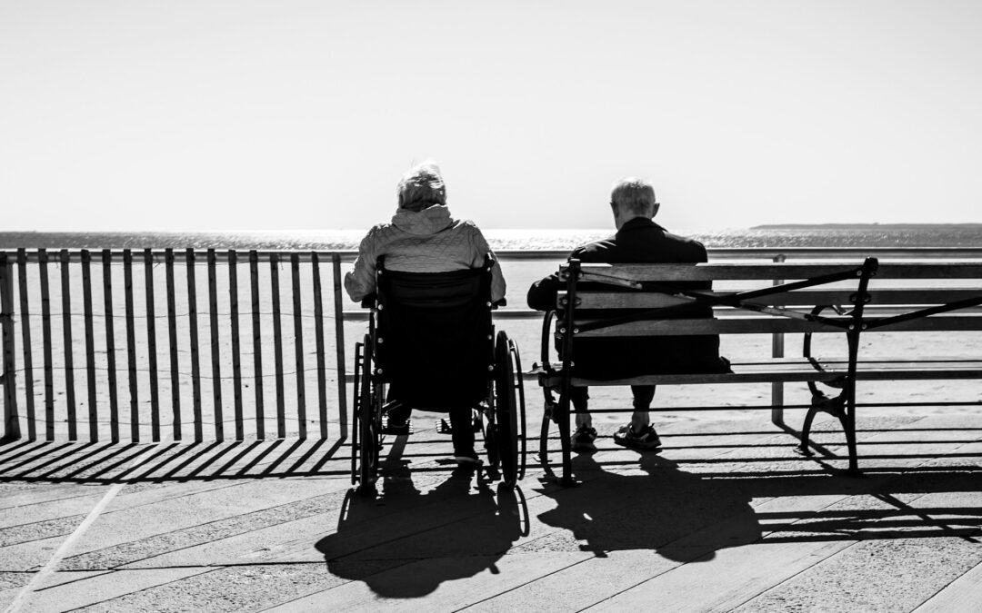 An elderly couple photographed from behind, sitting on the waterfront looking out to sea. She is in a wheelchair, he is sitting beside her on a bench seat.