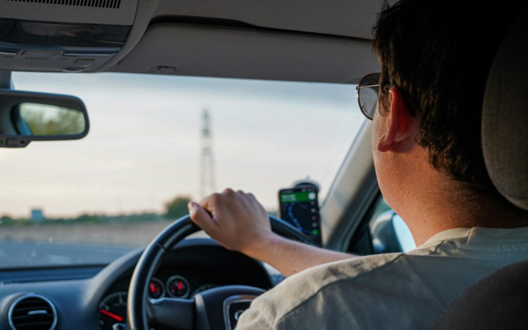 Side view of a man driving a car, shot taken from the rear left passenger seat. He has one hand on the wheel, is wearing sunglasses and has a mobile phone mounted on the dash board showing directions.