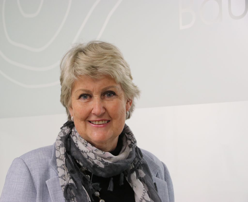 Practice Manager standing at reception. Wearing a black jacket, white top and silver necklace. Reception desk has a small green indoor plant sitting on it.