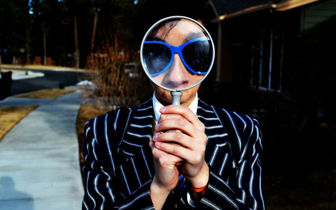 Male holding large magnifying glass in front of his face. Wearing large blue sun glasses and a stiped blazer.